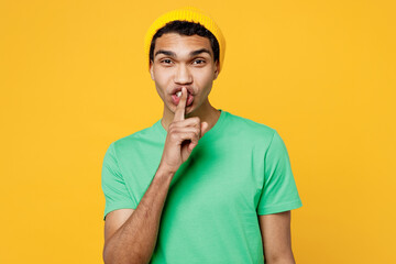 Young man of African American ethnicity wear casual clothes green t-shirt hat say hush be quiet with finger on lips shhh gesture isolated on plain yellow background studio portrait. Lifestyle concept