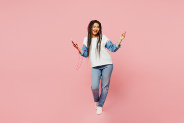 Full body young woman of African American ethnicity in white sweatshirt casual clothes headphones listen to music use mobile cell phone isolated on plain pastel light pink background studio portrait