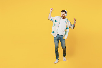 Full body young excited man he wear blue shirt white t-shirt casual clothes listen to music in headphones raise up hands dance isolated on plain yellow background studio portrait. Lifestyle concept.