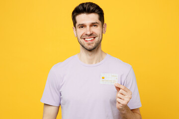 Young happy smiling fun man he wearing light purple t-shirt casual clothes hold in hand mock up of credit bank card look camera isolated on plain yellow background studio portrait. Lifestyle concept.