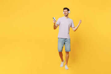 Fototapeta na wymiar Full body young smiling caucasian man wears light purple t-shirt casual clothes headphones listen to music use mobile cell phone isolated on plain yellow background studio portrait. Lifestyle concept.