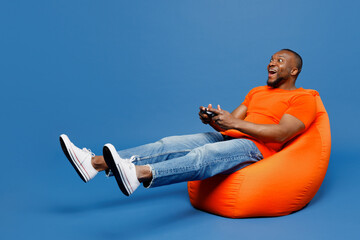 Full body young man of African American ethnicity in orange t-shirt sit in bag chair hold in hand...