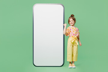 Full body fun little child kid girl 6-7 years old wear casual clothes point finger on big huge blank screen area mobile cell phone isolated on plain green background. Mother's Day love family concept.