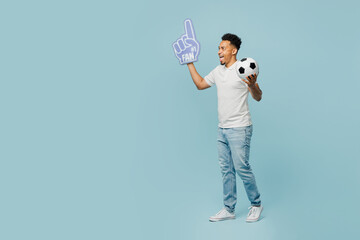 Full body side view young man fan wearing basic t-shirt foam 1 fan glove finger up cheer up support football sport team hold soccer ball watch tv live stream isolated on plain blue color background.