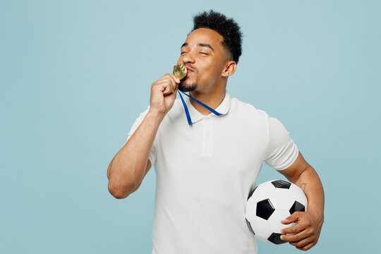 Young winner happy man fan wears basic t-shirt kissing golden medal cheer up support football sport team hold in hand soccer ball watch tv live stream isolated on plain pastel blue color background.
