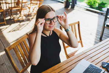 Female manager, freelancer, enjoying coffee break on summer terrace of cozy cafe, working remotely on laptop, smiling looking at laptop. Work online in the summer