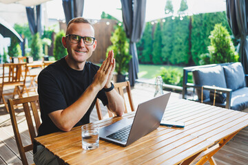 A successful male freelancer in a black t-shirt and glasses sits on the summer terrace of a cafe and works on a laptop. The manager sits in a cafe during lunch and works online.