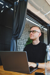 A successful male freelancer in a black t-shirt and glasses sits on the summer terrace of a cafe and works on a laptop. The manager sits in a cafe during lunch and works online.