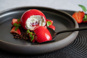 Selective focus of a piece of sphere curd cake on the black spoon. Strawberries and brownies. Dessert with red smooth surfaces and mirror glaze on the black plate.