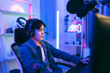 A young Asian man professional gamer wearing jeans jacket sits on a chair with a gaming table with...