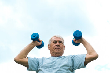 Senior man working out outdoor. Person lifting dumbbells. Old male exercising against blue sky....