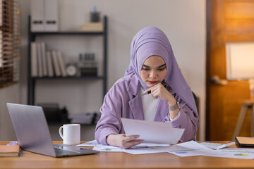 Fototapeta premium A stressed Asian business asian islamic woman in hijab holding documents is working from home, feeling stressed because of lot of paperwork appearing worried, tired, and overwhelmed