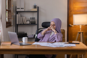 A stressed Asian business asian islamic woman in hijab holding documents is working from home, feeling stressed because of lot of paperwork appearing worried, tired, and overwhelmed