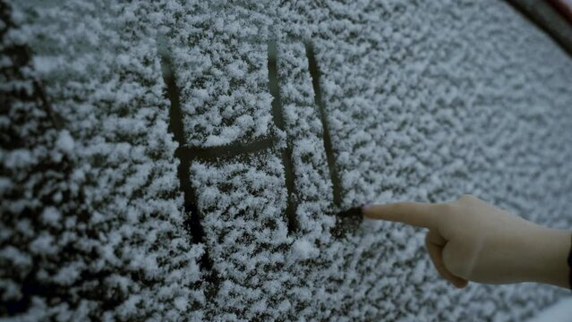 The girl writes with her finger on the snow-covered glass of the car the inscription - help. Close-up.
