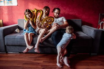 Little girl in festive cap stuck head into the foil baloon ONE. Portrait of happy, diversity family that fools around, having fun for year old baby. Birthday party at home. Lifestyle atmosphere