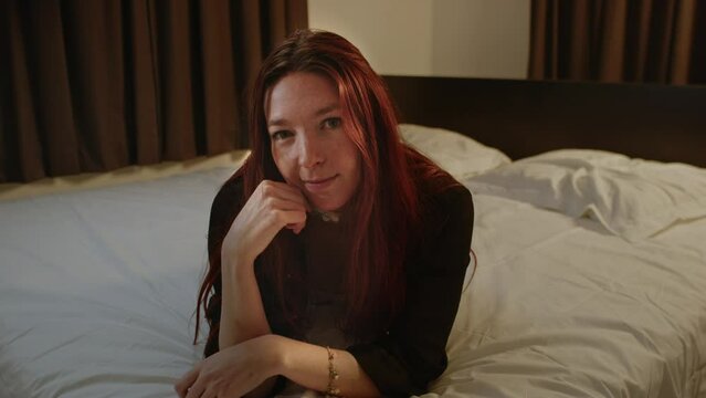 A beautiful red haired girl lies in bed and scratches her head while looking at the camera