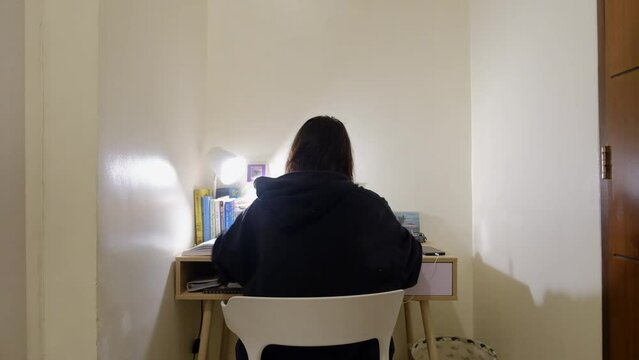 Static wide shot of girl with black hair in jacket sitting and studying at desk.