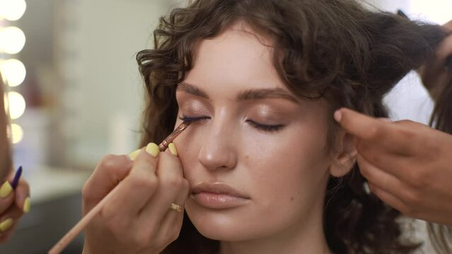 Close-up portrait of sensual curly woman being given from professional make-up artist and hairdresser doing makeup and hairstyle in beauty salon. Concept of four-handed makeup and styling.