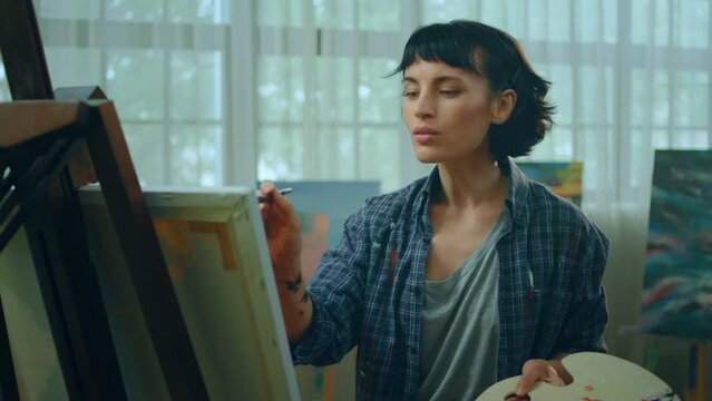 A woman seems very focused on her work as she is painting something on a canvas and wearing a blue flannel shirt with paint stains all over it