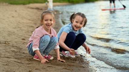 Little girls look at camera and laugh playing with sand on river bank in summer park at sunset. Childrens leisure games outdoors by water