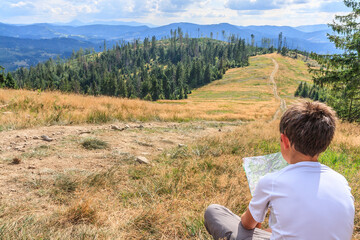 Fototapeta na wymiar A boy tourist with a map in his hand watching the panorama of the Beskid Żywiecki peaks (Poland) from the viewpoint on the yellow tourist trail under Hala Redykalna on a sunny summer day