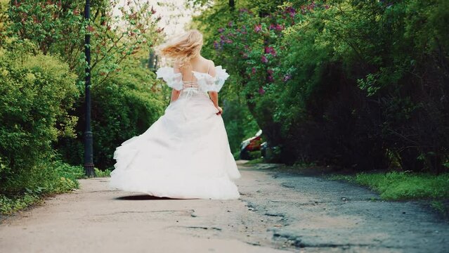 happy woman running on road waiting love, long blonde hair white boho style dress train fly in wind. Cute fantasy girl princess bride back rear view. Summer nature green leaves trees wedding day 4k