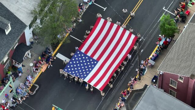 A small crowd watches a large Flag is carried past as part of the 4th July parade. Aerial view.