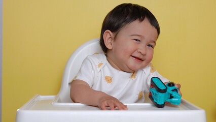 Cute asian baby sits on highchair with toy and smiling on yellow background