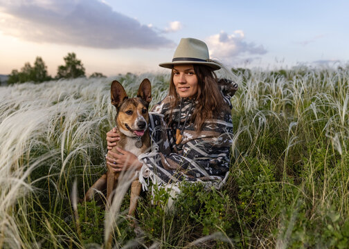 Portrait of a woman in a poncho and a hat with her beloved dog. Summer landscape, feather grass field, sky with clouds.