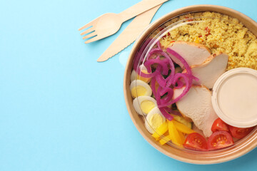 Tasty food in container with wooden fork and knife on light blue background, flat lay. Space for text