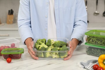 Man holding glass container with fresh broccoli at white marble table in kitchen, closeup. Food storage