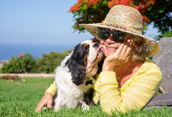 Senior smiling woman lying down in the meadow with her cavalier king charles spaniel dog kissing her. Elderly lady and her best friend