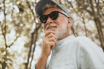 Portrait of senior bearded man with black sunglasses touching his beard looking away smiling enjoying environment in trekking day in mountain forest