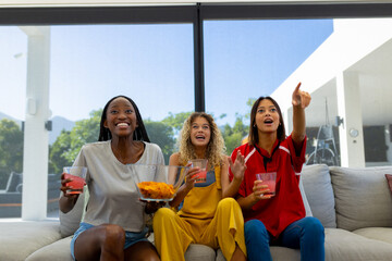 Group of excited diverse friends sitting on sofa, watching sport on tv and pointing