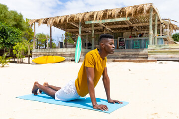 Focused biracial man practicing yoga, stretching on mat on sunny beach, unaltered