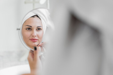 A beauty-loving woman wearing a towel with a healthy, smooth body looking at a round mirror in a white bathtub in a bathroom..