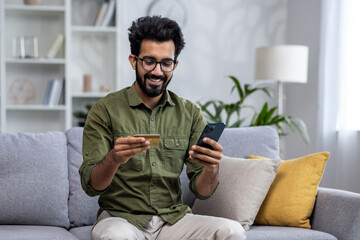 Happy and satisfied man shopping online sitting on sofa at home, hispanic customer holding bank...