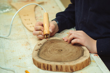 Kid makes wooden clock in the workshop. Boy burn out numbers with soldering iron on wooden disc.