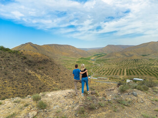Obraz premium Young couple of travelers in love stands on the edge of a mountain and looks into the distance at a garden of trees surrounded by brown mountains, view from a drone. Concept of travel, tourism.