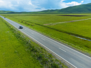 Black passenger car is driving along an asphalt road along green fields, mountains on a sunny day. Photographing from a drone. Concept of travel