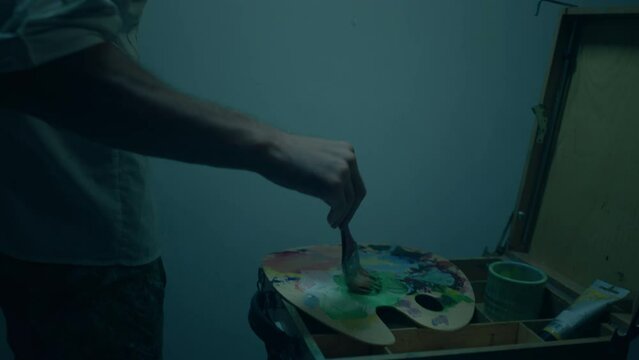 In the dark art studio charismatic man artist take the paint brush and mixing some oil colour from the pallet and add on the wall he painting new contemporary picture