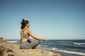 Fototapeta na wymiar a beautiful athletic brunette girl in a gray tracksuit is engaged in meditation sitting on the sand against the backdrop of the sea