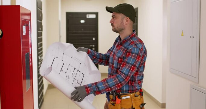 A worker stands in the corridor of a multi-storey building, holds a paper plan in his hands and looks at the fire shield. Checking fire safety boards.
