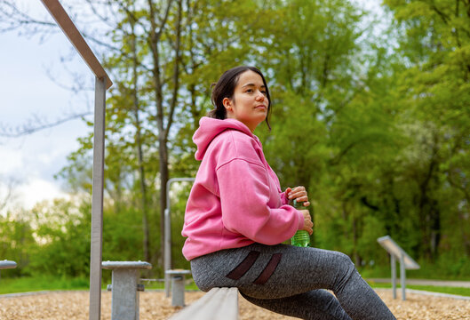 young woman in outside outdoor gym using cable chest for workout or parallel bars.girl female is resting sitting down drinking water from plastic bottle.pink hoodie park environment