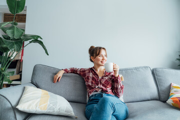 Young smiling woman sitting on sofa and looking away while drinking coffee or tea. Young brunette woman relaxing after housekeeping, home cleaning. Portrait of relaxed female resting at home. - Powered by Adobe