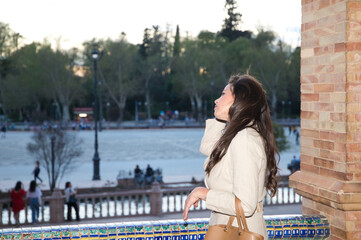 Young and beautiful woman with long brown hair is in the most famous square in Seville. The woman looks out from a balcony of the monument to see the centre of the square. The woman is happy