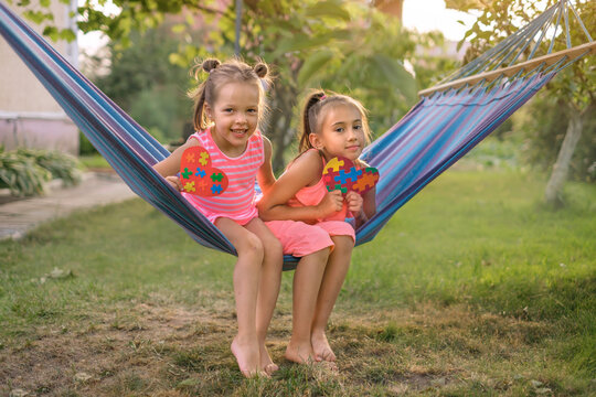 Charming hilarious sisters have fun in the yard on a hanging hammock on a warm summer day . Children hold in their hands earrings made of multi-colored puzzles - Powered by Adobe