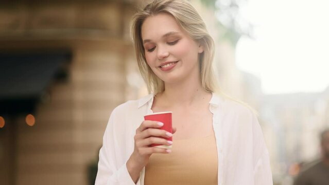 Close up portrait of charming blond woman enjoying cup of coffee or tea outdoors Pretty smiling girl relaxing at city street alone Beautiful day concept