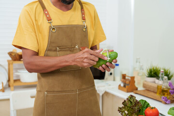 Bearded Asian man in overalls standing in the kitchen He is using his hand to peel a mango that is...