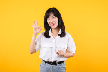 Young businesswoman hand sign okay gesture. Asian woman happy smile wearing white shirt and jean denim plants showing okay sign hand gesture isolated on yellow background.